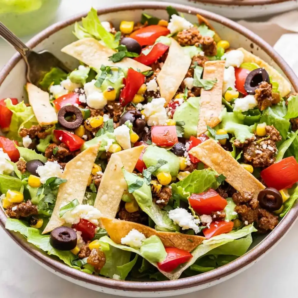 Taco Salad Recipe: Crispy Beef and Cool, Crunchy Layers in 32 Minutes 7 Taco salad with seasoned beef, lettuce, corn, tomatoes, black olives, tortilla strips, cheese, and creamy avocado dressing.