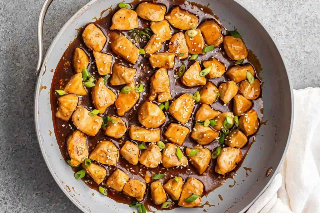 Teriyaki chicken pieces simmering in a skillet with glossy sauce, sesame seeds, and sliced green onions.